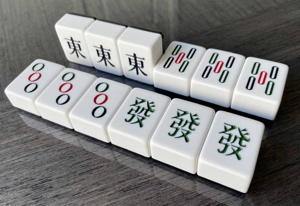 A set of Mahjong tiles arranged in matching patterns, featuring East wind, Bamboo, Circles, and Green Dragon tiles, displayed on a glossy surface.